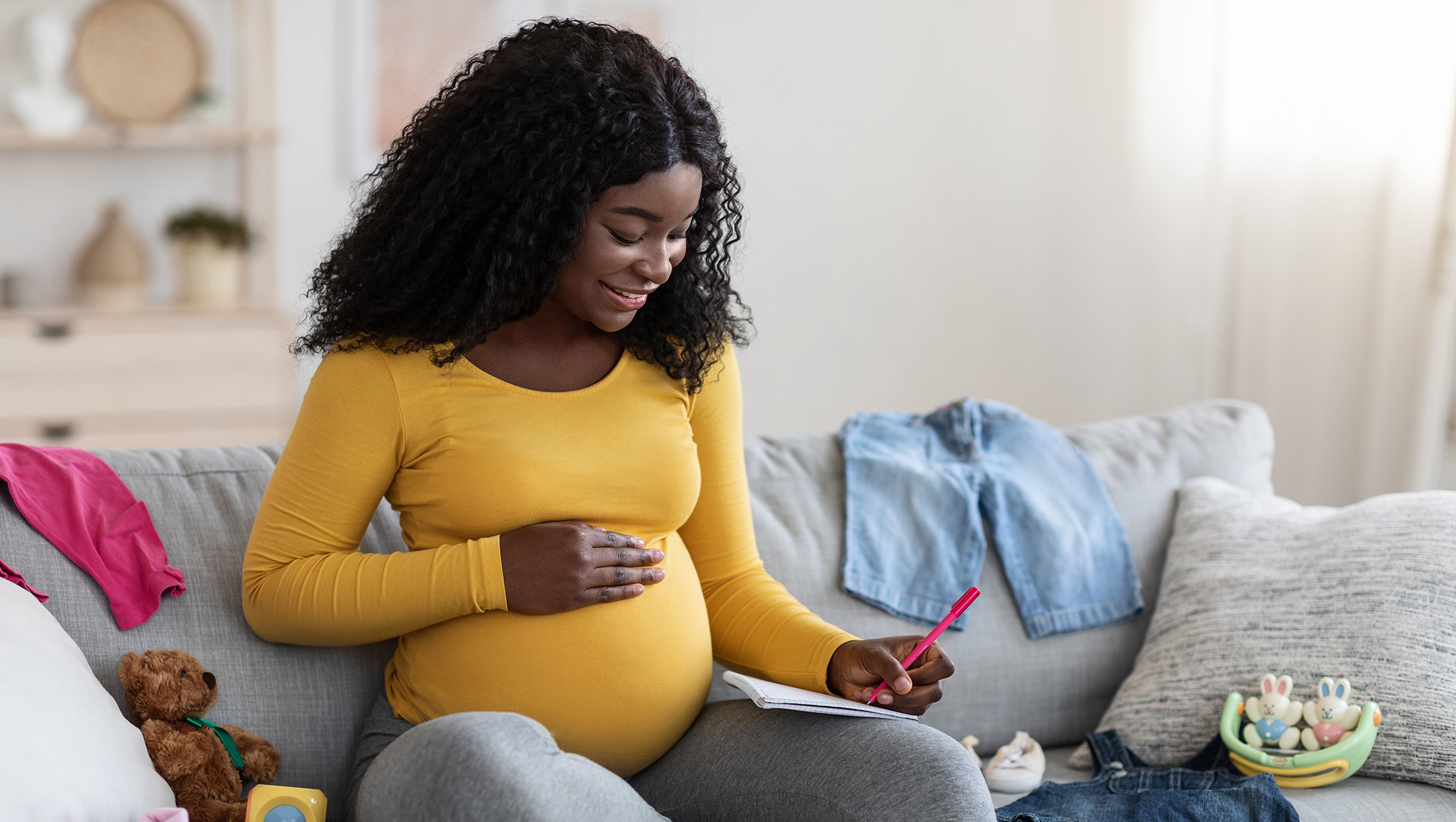 A happy pregnant woman sits on her sofa with a notebook and pen.